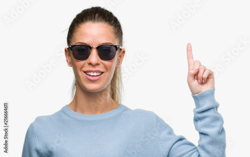 Beautiful young woman wearing sunglasses and ponytail surprised with an idea or question pointing finger with happy face, number one