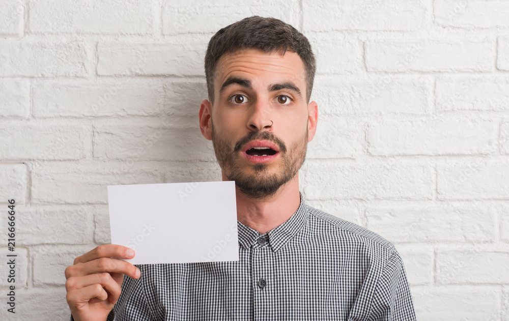 Young adult man over brick wall holding blank paper scared in shock with a surprise face, afraid and excited with fear expression Young adult man over brick wall holding blank paper scared in shock with a surprise face, afraid and excited with fear expression