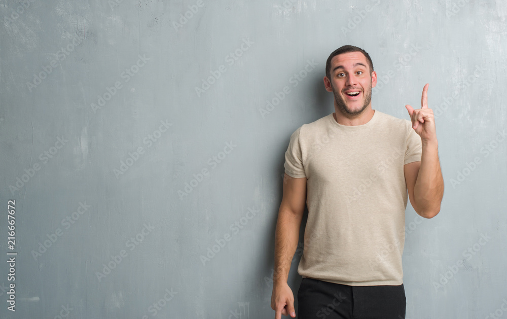 Young caucasian man over grey grunge wall pointing finger up with successful idea. Exited and happy. Number one. Young caucasian man over grey grunge wall pointing finger up with successful idea. Exited and happy. Number one.