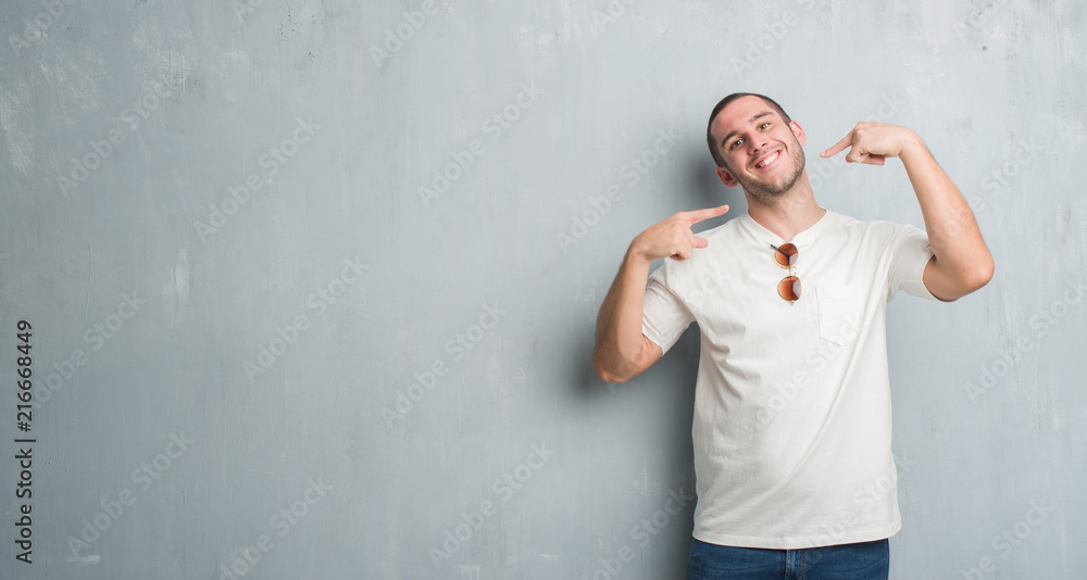 Young caucasian man over grey grunge wall wearing sunglasses smiling confident showing and pointing with fingers teeth and mouth. Health concept.