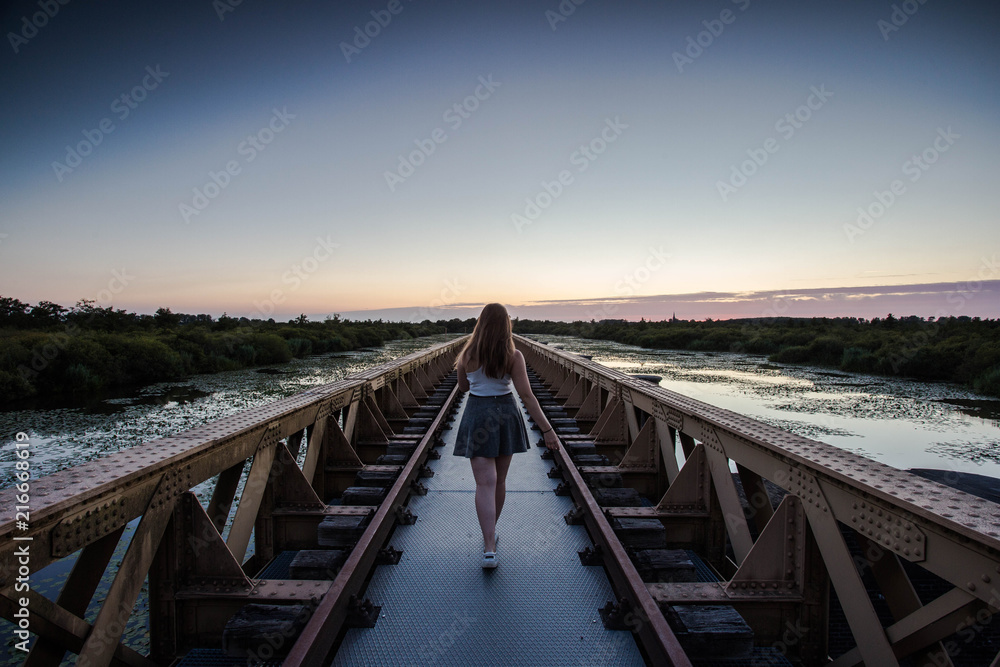 A happy young slender girl walks near a bridge in the rays of the summer sun.