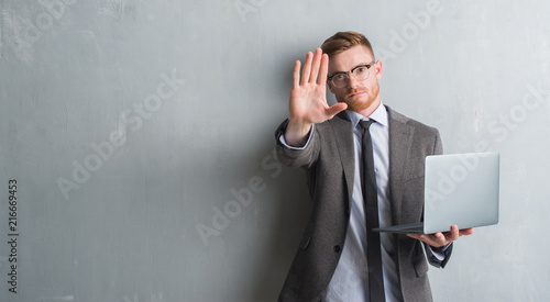 Young redhead elegant business man over grey grunge wall using laptop with open hand doing stop sign with serious and confident expression, defense gesture
