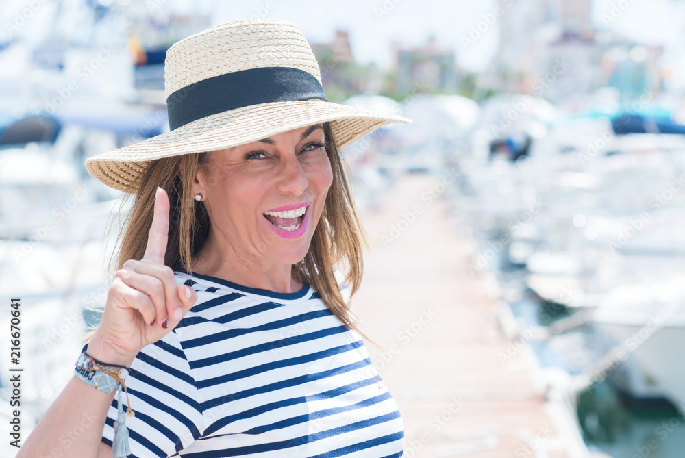 Beautiful middle age woman standing in marina surprised with an idea or question pointing finger with happy face, number one