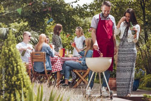 Smiling woman drinking beer while her friend grilling food during birthday outdoor party