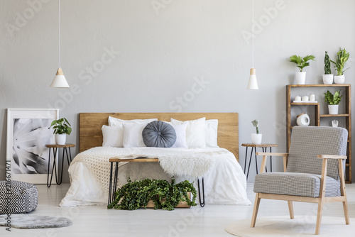 Patterned armchair and plants in bright bedroom interior with poster next to wooden bed. Real photo