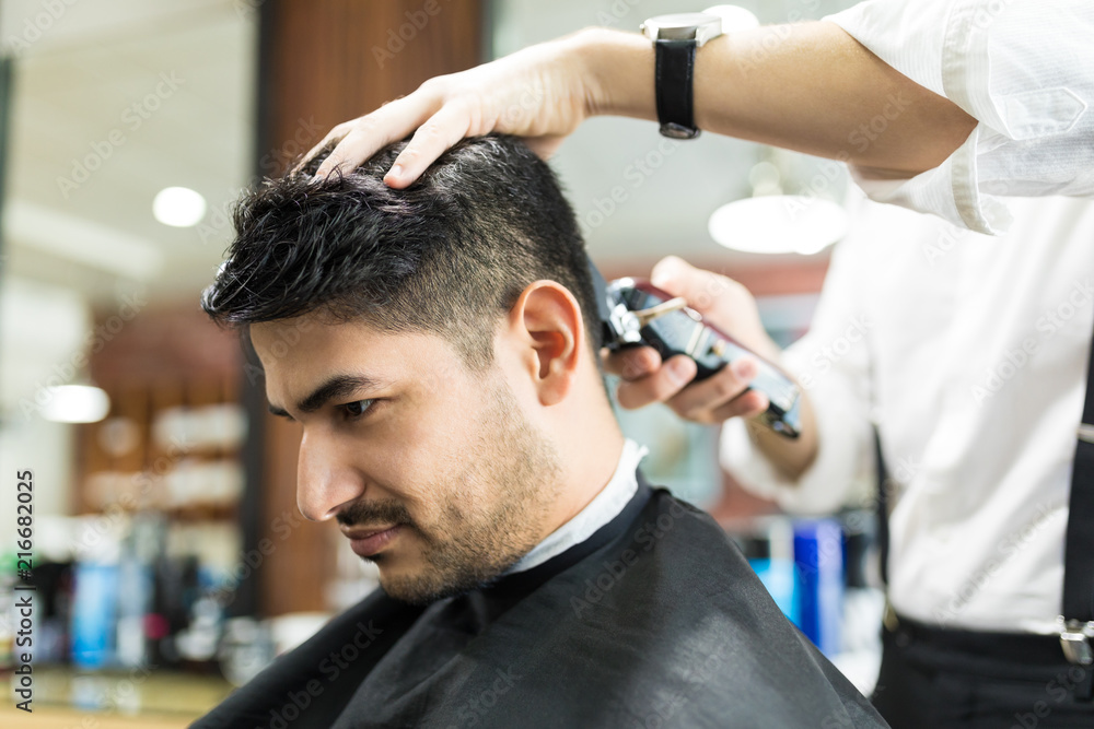 Fototapeta premium Male Receiving Haircut By Barber Using Trimmer In Salon