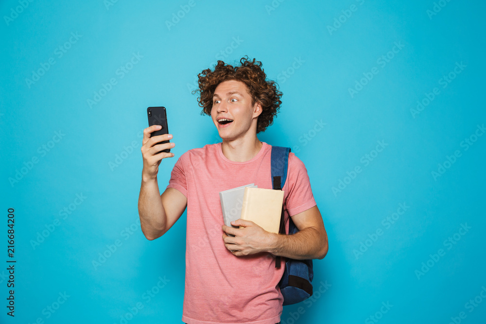 Image of attractive youngster guy with curly hair wearing casual clothing and backpack holding smartphone and books, isolated over blue background Image of attractive youngster guy with curly hair wearing casual clothing and backpack holding smartphone and books, isolated over blue background