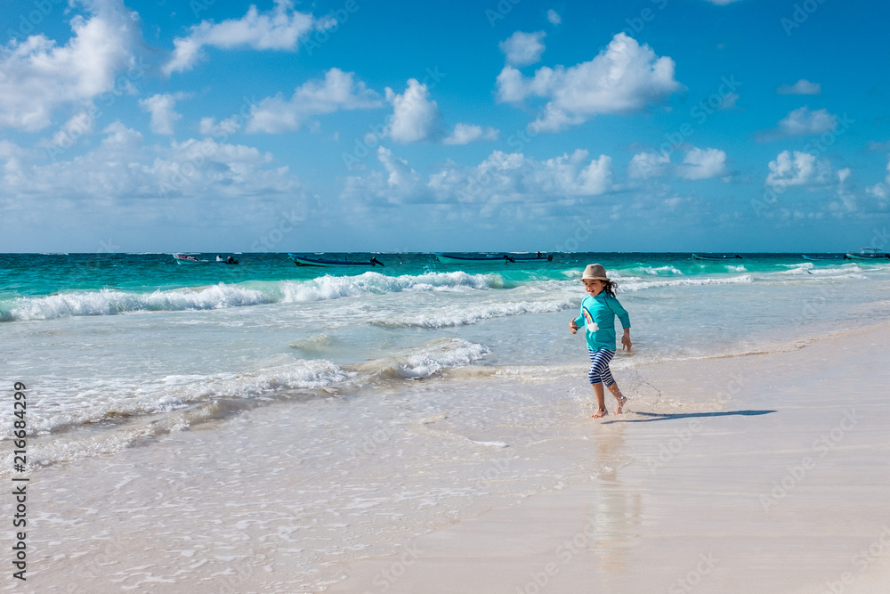 Little girl wearing a sunhat running down the beach with small waves in the background