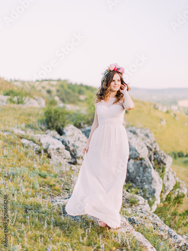 The bride is touching the curly hais and looking aside while standing on the mountains. photo