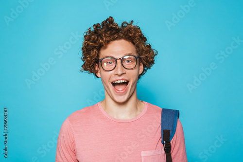 Image of geek guy with curly hair wearing glasses and backpack smiling at camera, isolated over blue background photo