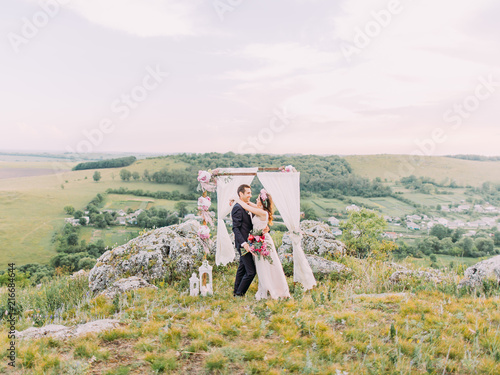 Horizontal photo of the hugging newlywed couple near the lovely decorated wedding arch placed in the mountains. photo