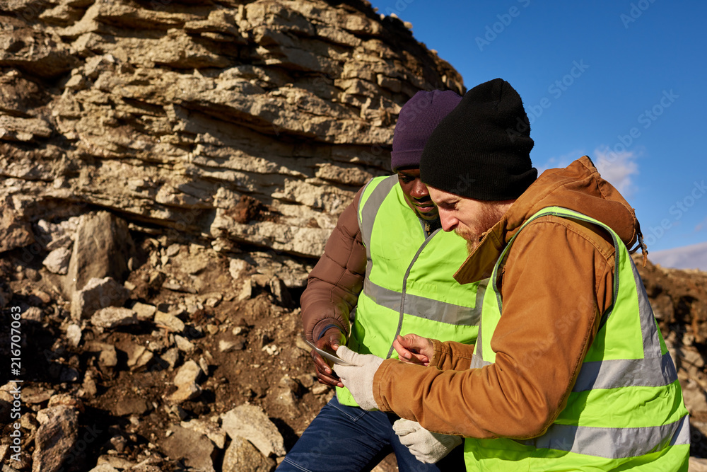 Naklejka premium Side view portrait of two industrial workers wearing reflective jackets, one of them African, inspecting mineral mines on worksite outdoors and using digital tablet, copy space