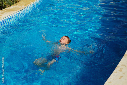 Happy kid playing in blue water of swimming pool on a tropical resort at the sea. Summer vacations concept. Cute boy swimming in pool water