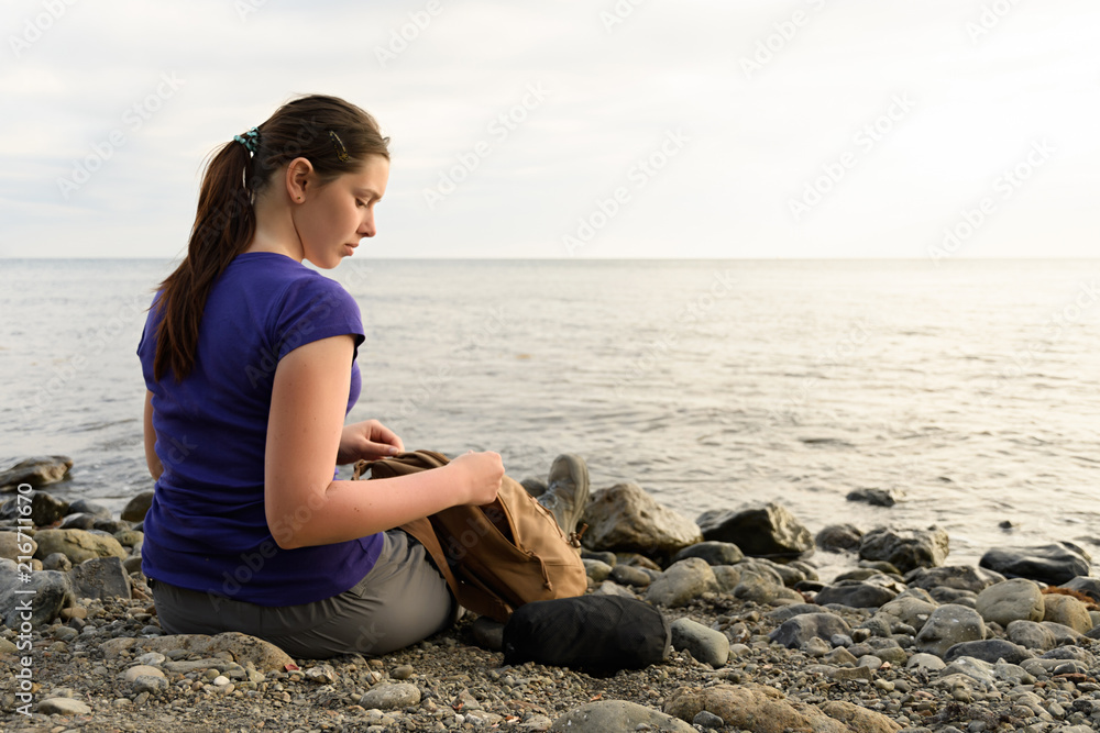 Tourist girl sitting on the beach and pulls things out of the backpack on the shore
