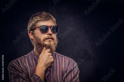 Close-up portrait of a pensive bearded redhead guy wearing sunglasses dressed in an old-fashioned shirt on a dark background.