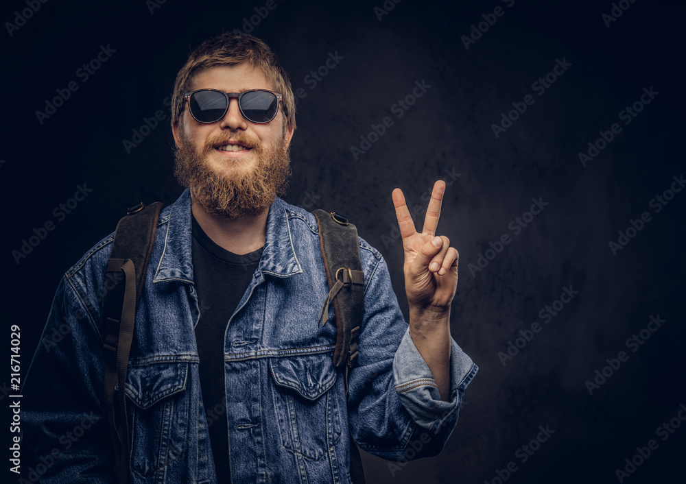 Smiling bearded hipster guy wearing sunglasses and backpack dressed in jeans jacket showing peace gesture. Isolated on a dark background. Smiling bearded hipster guy wearing sunglasses and backpack dressed in jeans jacket showing peace gesture. Isolated on a dark background.