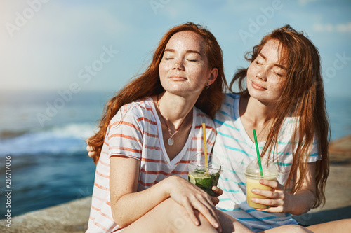 Beautiful redhead girls enjoying warm sunlight, closing eyes and resting near sea, drinking cocktails, hugging and closing eyes to relax, care bad thoughts away, feeling carefree on summer vacation