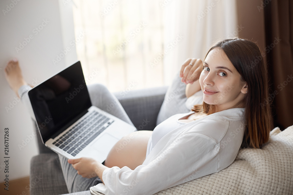 Pregnant woman chilling and relaxing at home, asking husband glass of milk while watching video about maternity, lying on armchair and holding laptop on laps, smiling with cute expression at camera Pregnant woman chilling and relaxing at home, asking husband glass of milk while watching video about maternity, lying on armchair and holding laptop on laps, smiling with cute expression at camera