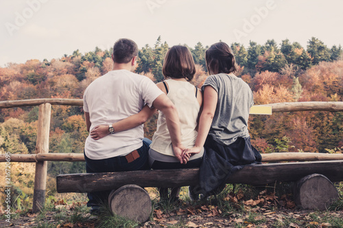 Three young beautiful people sit on a wooden bench and enjoy the autumn forest tree landscape, the concept of a love triangle, betrayal and treachery