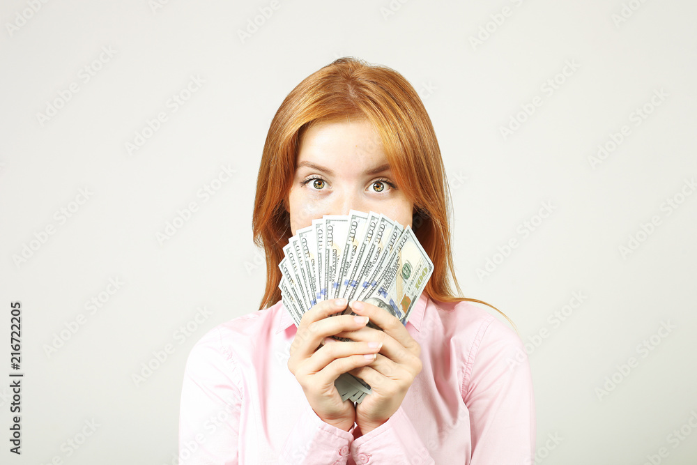 Young beautiful redhead woman covering her face with fistful of money holding bunch of one hundred dollar bills like fan. Excited attractive female with lots of cash. Background, copy space, close up. Young beautiful redhead woman covering her face with fistful of money holding bunch of one hundred dollar bills like fan. Excited attractive female with lots of cash. Background, copy space, close up.