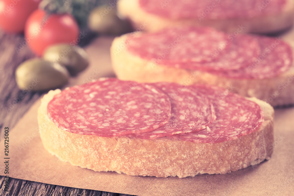 Salami slices on ciabatta bread with green olives and cherry tomato in the back (Selective Focus, Focus on the front of salami slices on first sandwich) (Digitally Altered: Toned Image) Salami slices on ciabatta bread with green olives and cherry tomato in the back (Selective Focus, Focus on the front of salami slices on first sandwich) (Digitally Altered: Toned Image)