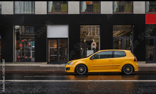 Yellow car is parked at asphalt road in the city center in the rain. Storefront