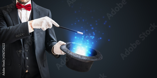 Tela Magician or illusionist is showing magic trick with wand and hat on dark background