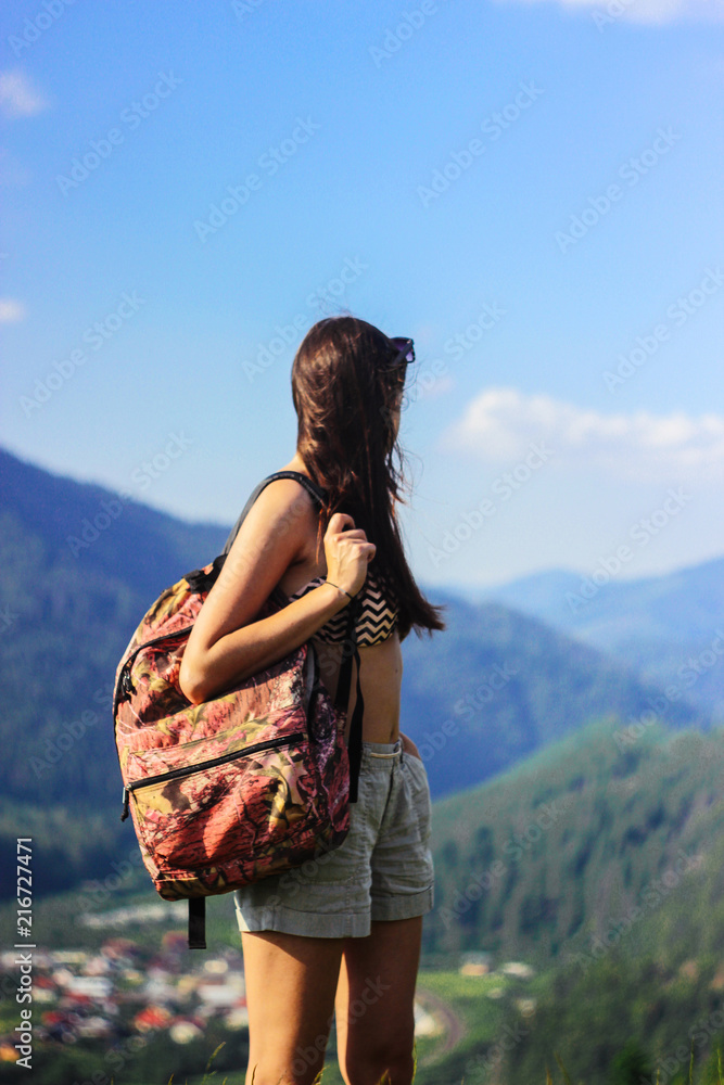 happy beautiful girl hipster with backpack, looking at city down the hill on background of amazing sky and mountains in summer