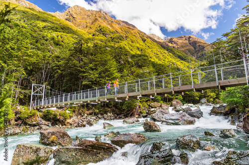 New Zealand tramping people crossing river bridge. Hikers couple backpackers walking hiking together with backpacks on Routeburn Track trail path. photo