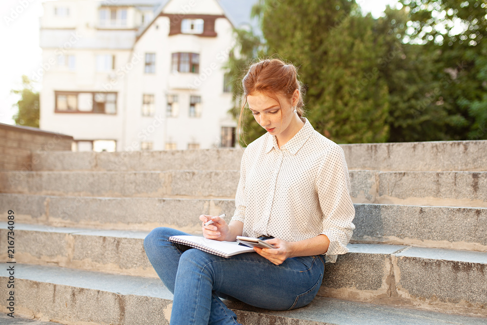 Young beautiful red hair girl with freckles sitting on a stairs near the university writing in a notebook with homework. Portrait of a student. Back to school concept. Young beautiful red hair girl with freckles sitting on a stairs near the university writing in a notebook with homework. Portrait of a student. Back to school concept.