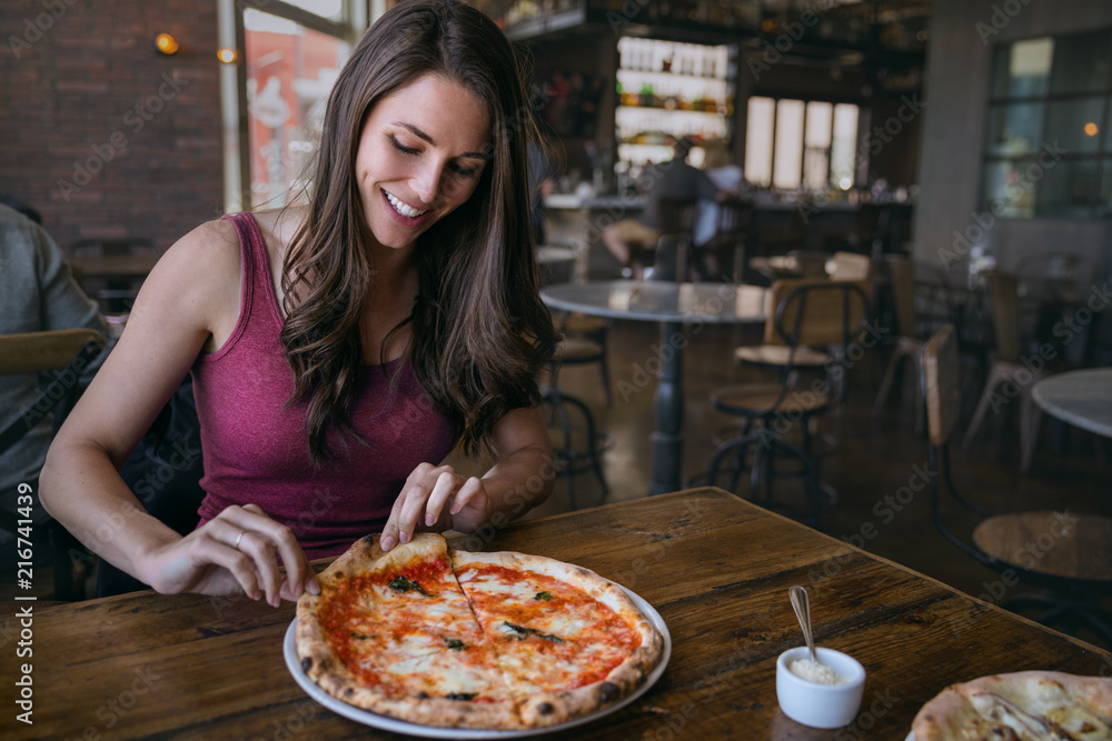Fototapeta Uśmiechnięta kobieta w modnej pizzerii rzemieślniczej, chwytając kawałek pysznej pizzy rzemieślniczej