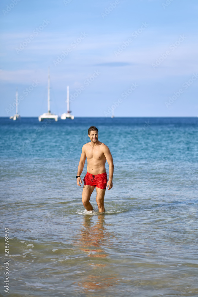 Fototapeta premium Tanned guy in red shorts on beach. Daylight shooting.