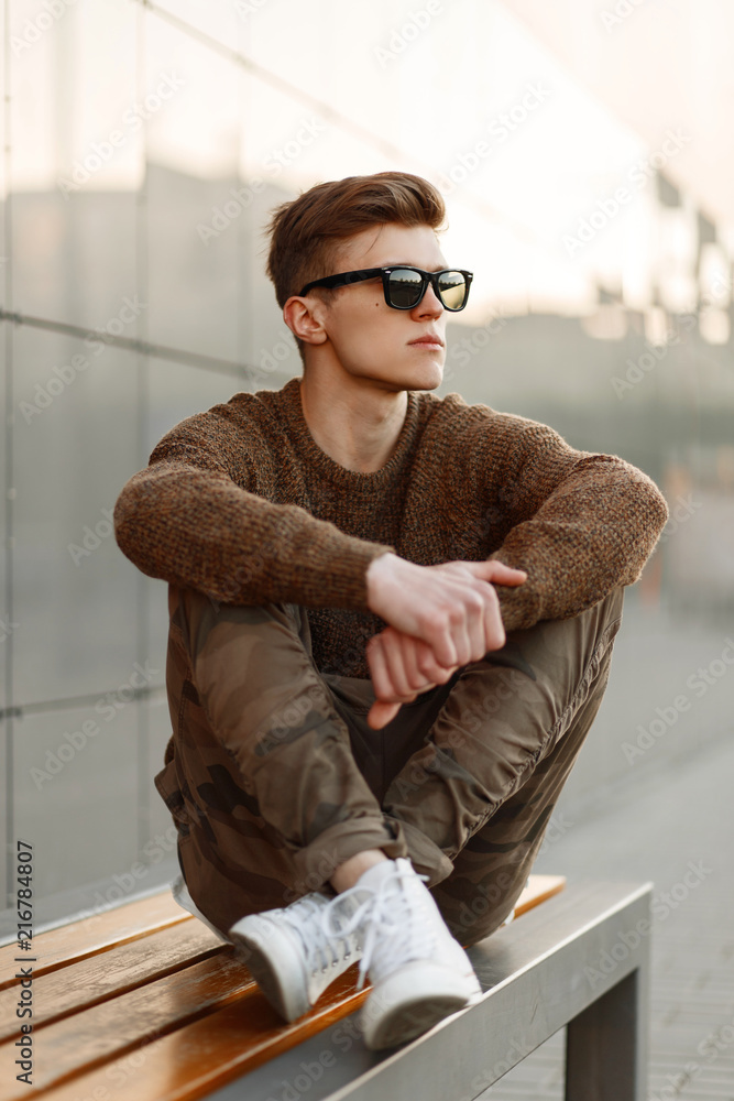 Stylish handsome man in sunglasses in a sweater and military fashion pants  with white sneakers sits on a bench in the city Stock Photo | Adobe Stock