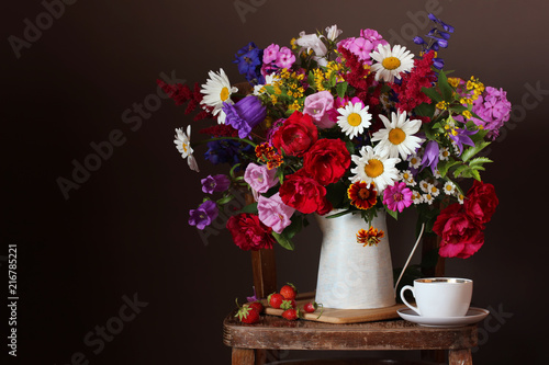 Bouquet of cultivated flowers in a jug on a chair and strawberries.