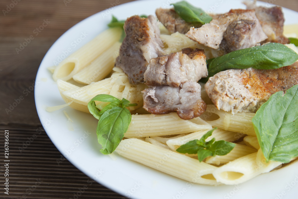 Cooked penne pasta isolated in white round plate, with basil leaves and pork meat, over wooden table, decorated with basil leaves. Top view. Cooked penne pasta isolated in white round plate, with basil leaves and pork meat, over wooden table, decorated with basil leaves. Top view.