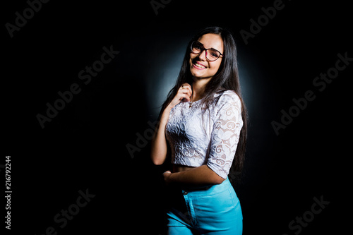 Portrait of an attractive young woman in white top and blue pants posing with her glasses in the dark.