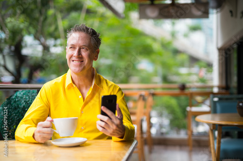 Mature Man Sitting In Coffee Shop And Thinking While Using Mobile Phone