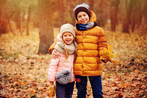 autumn portrait of happy kids playing outdoor in park. Smiling brother and sister walking in sunny day, wearing warm knitted hats and scarfs