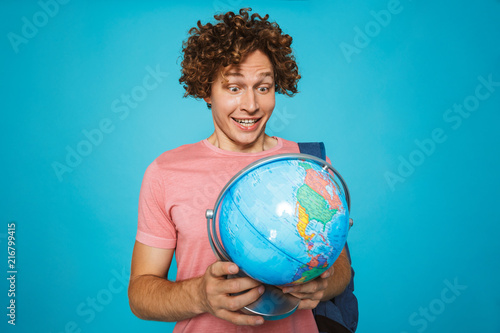 Portrait of european student guy with curly hair wearing backpack smiling and holding earth globe, isolated over blue background