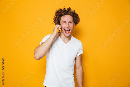 Photo of brunette joyous guy with curly hair talking on smartphone  isolated over yellow background