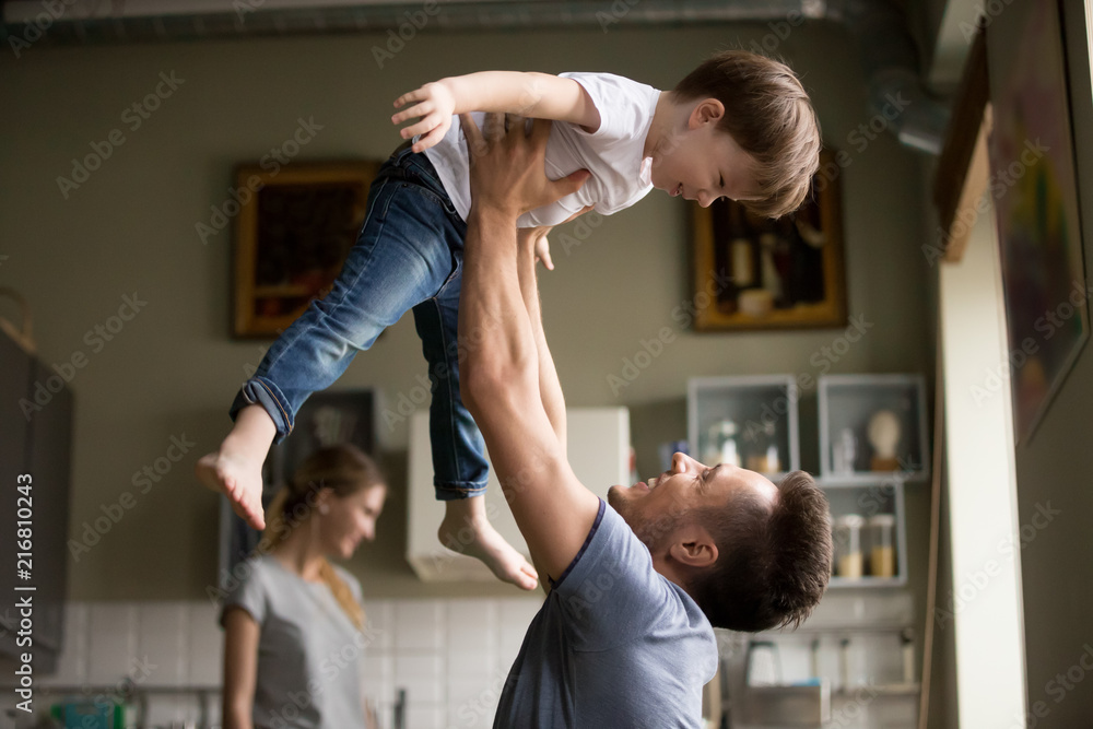 Fototapeta premium Happy dad lifting son up having fun together at home, young loving father and little child playing funny game in living room, daddy laughing enjoying spending time with kid boy and family on weekend