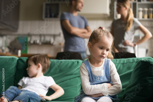 Upset little girl feeling sad after fight with brother sitting on sofa with worried parents on background, sulky frustrated sister ignoring child boy disinterested or bored, siblings rivalry concept photo