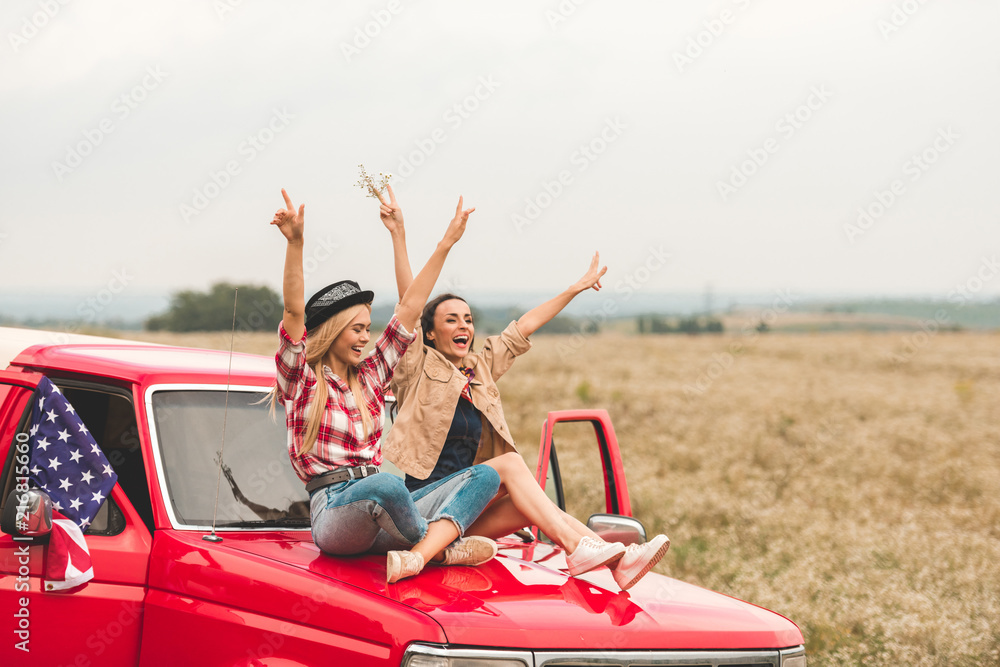 beautiful young girlfriends sitting on car hood with raised hands and showing peace signs