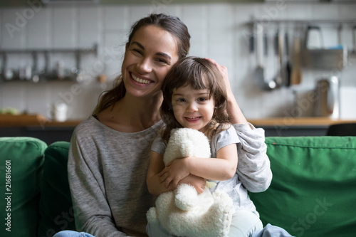 Portrait of happy family single mother and kid daughter embracing on sofa at home, young cheerful mom sister stroking cute girl hugging looking at camera, mum and child sincere relationship concept