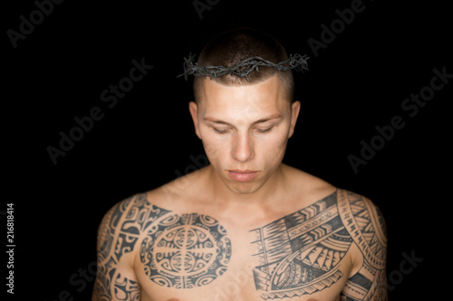 A guy with tattoos on the body topless in a wreath of wire with spikes on a black background