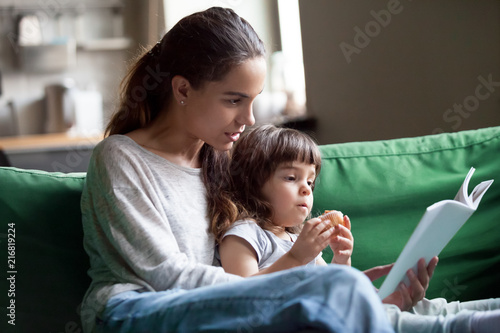 Canvastavla Young mother teaching daughter holding book sitting on sofa, mom or babysitter e