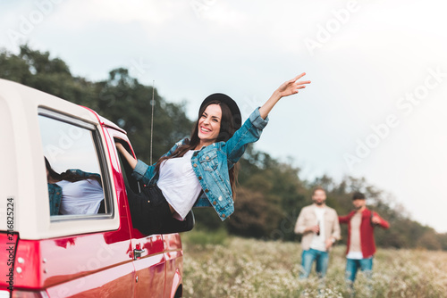 young woman outstretching from car window and raising hand while men standing blurred on background in flower field