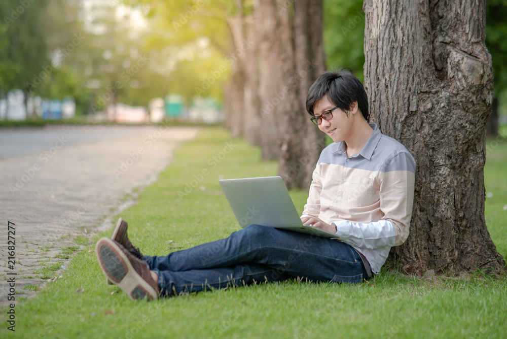 Young Asian happy man using laptop computer white sitting on grass in the park. Male university student relaxing outdoor. Education and technology concepts Young Asian happy man using laptop computer white sitting on grass in the park. Male university student relaxing outdoor. Education and technology concepts