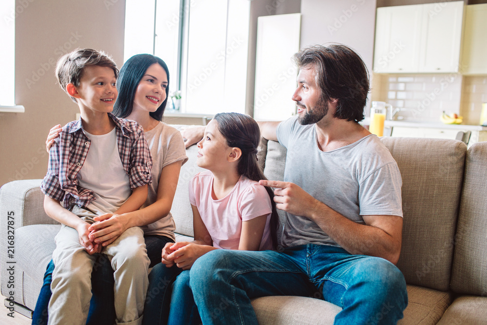 Happy family sits together on sofa. Boy is on mother's lap. Girl sits between woman and man. They are looking at each other. Happy family sits together on sofa. Boy is on mother's lap. Girl sits between woman and man. They are looking at each other.