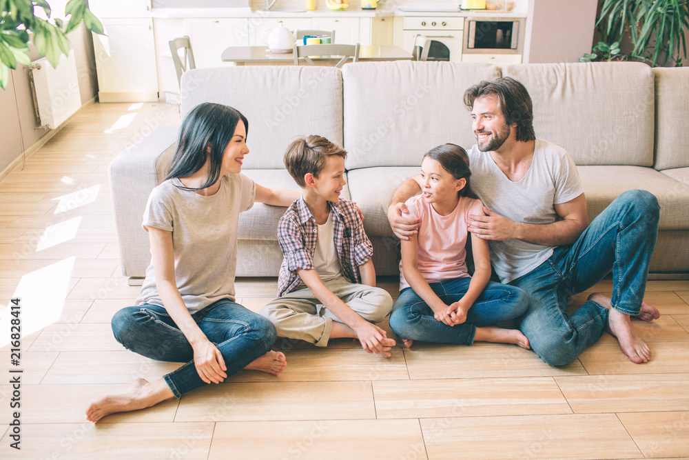 Positive and attractive people are sitting on the floor. They look at each other. Parents and kids wear jeans and shirts. They sit with legs crossed. Positive and attractive people are sitting on the floor. They look at each other. Parents and kids wear jeans and shirts. They sit with legs crossed.